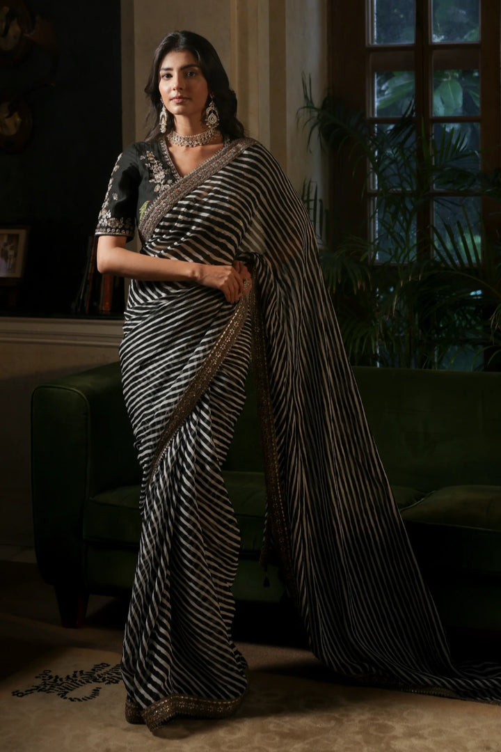 Woman in a black and white saree standing in a dimly lit room with large windows.