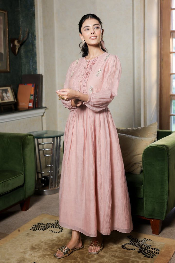 Woman in a pink dress standing in a living room with green furniture and a window in the background.