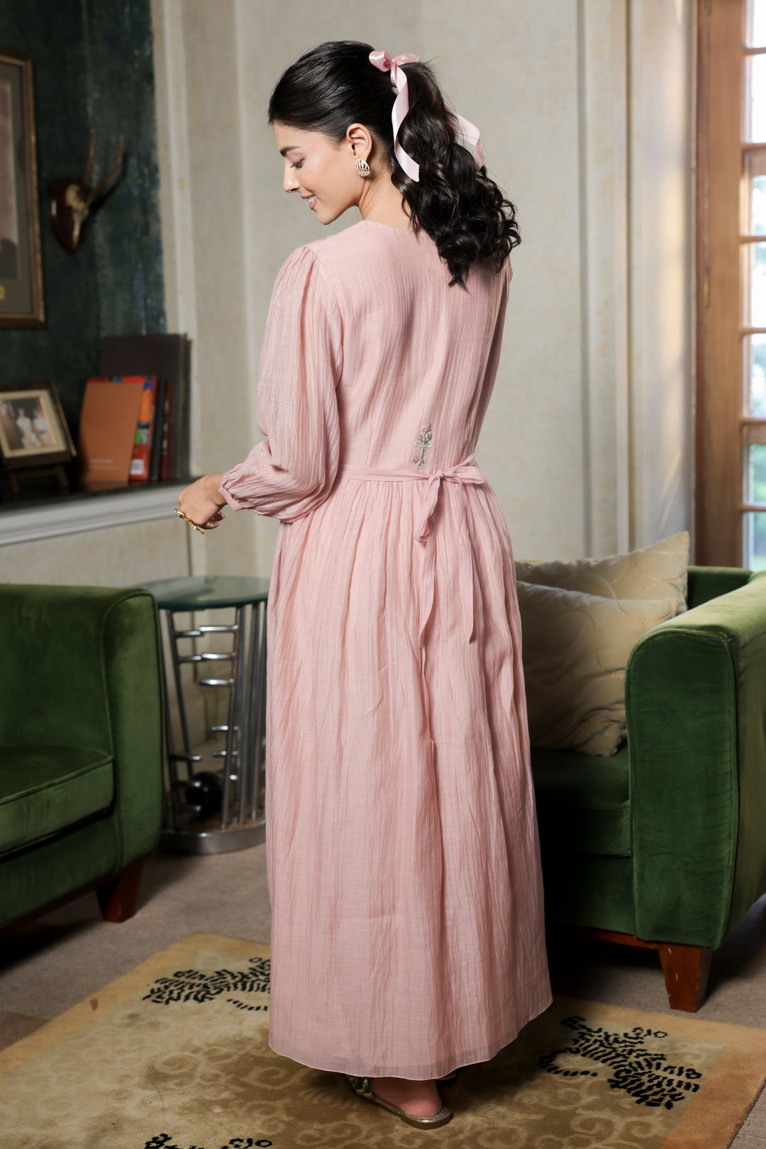 Woman in a pink robe standing in a living room with green furniture and a window.