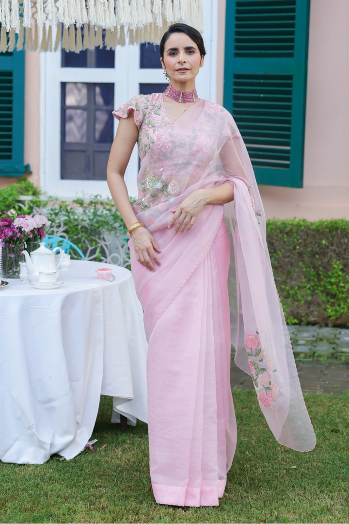 Woman in a pink saree standing outdoors with a decorative umbrella and table in the background.