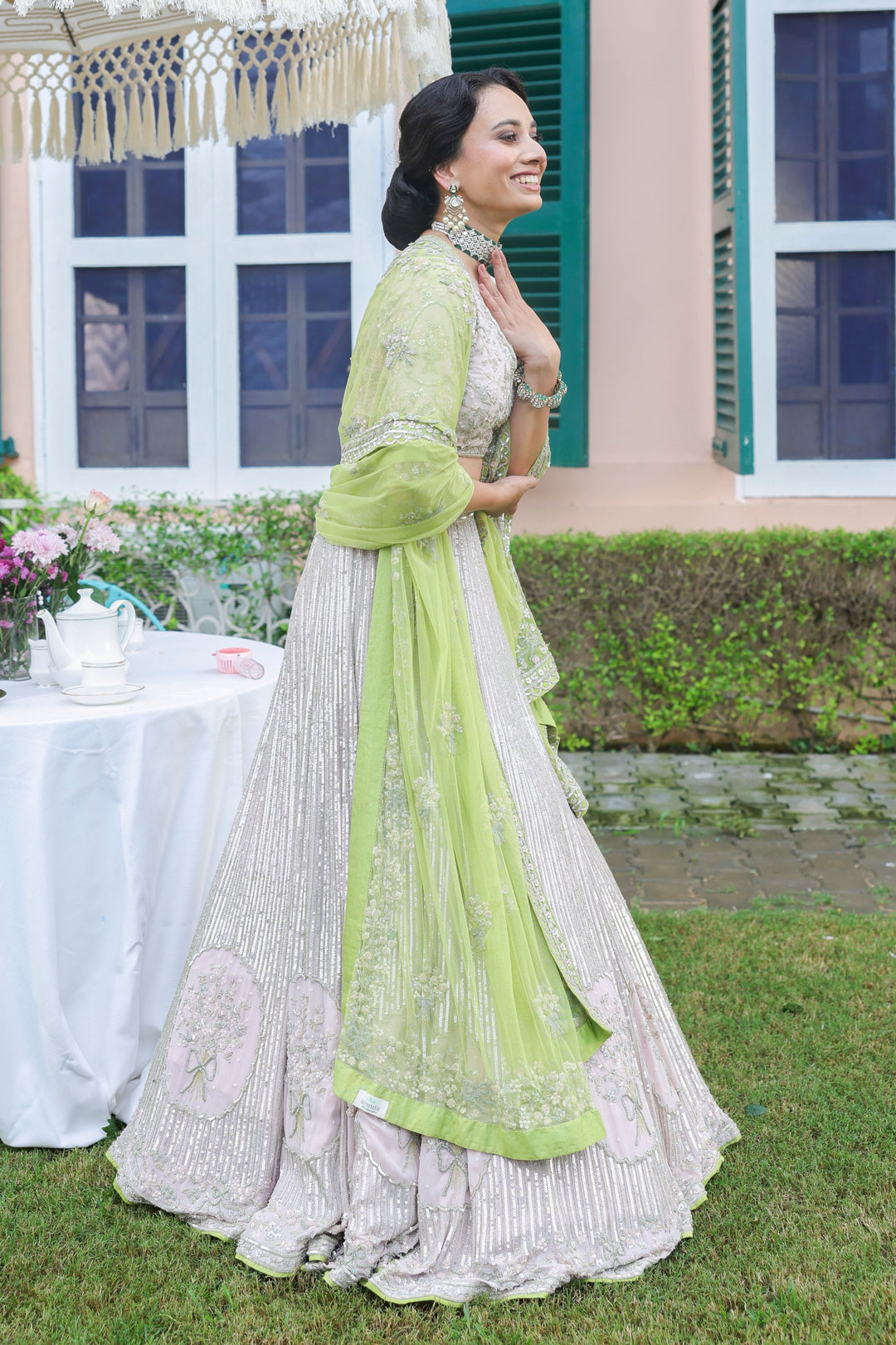 Woman in a light green and white traditional outfit standing outdoors with a table and umbrella in the background.