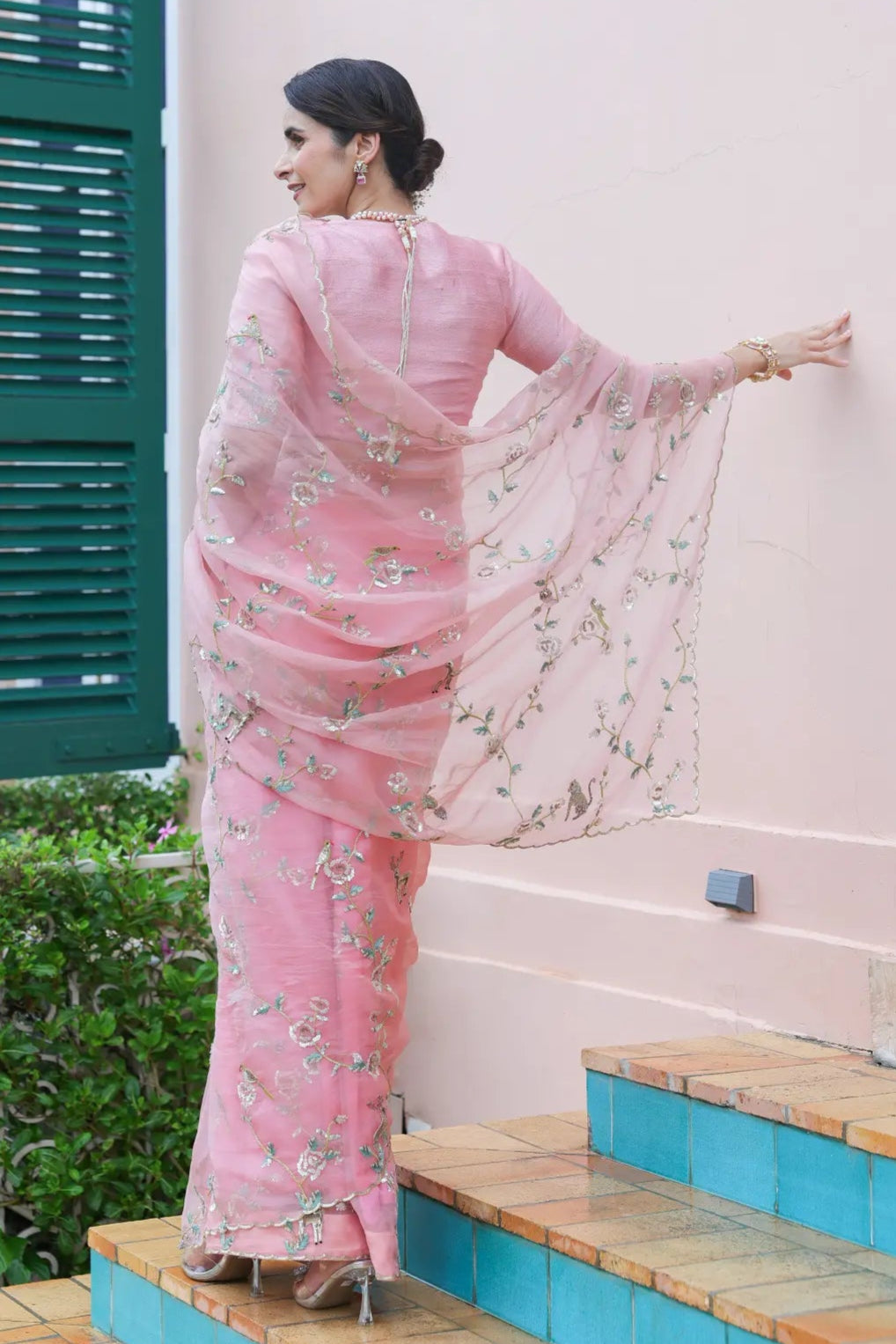 Woman in a pink saree standing on steps with a light pink wall and green shutters in the background