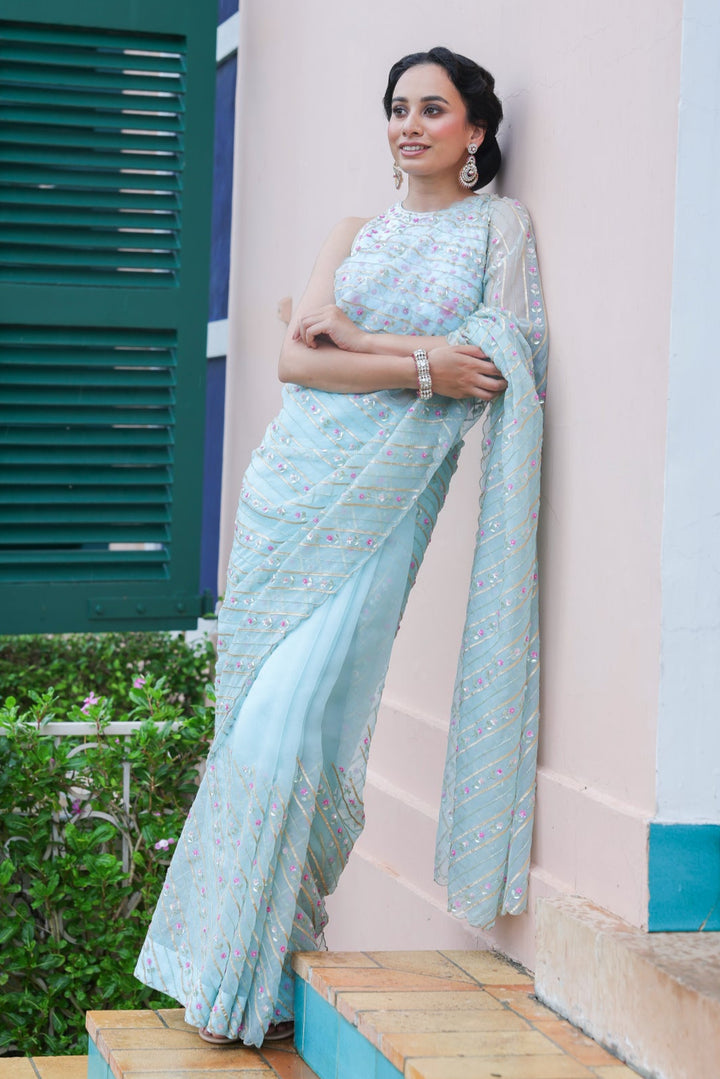 Woman in a light blue saree standing against a building with green shutters.