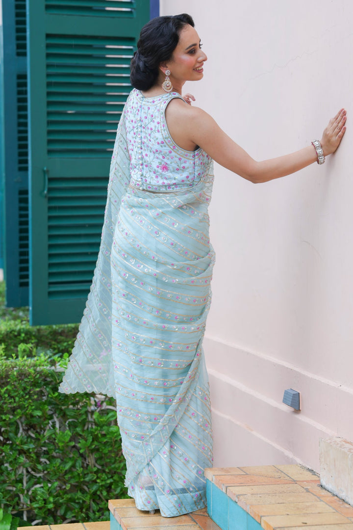 Woman in a light blue saree standing against a white wall with green shutters.