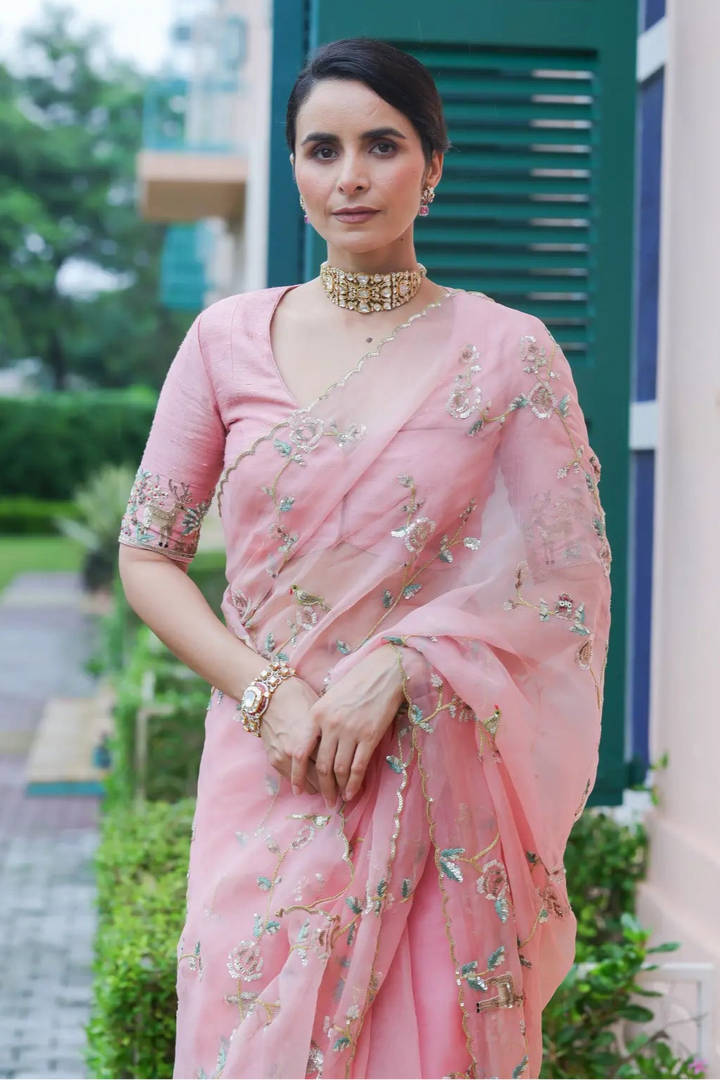 Woman in a pink saree standing outdoors with greenery and a building in the background
