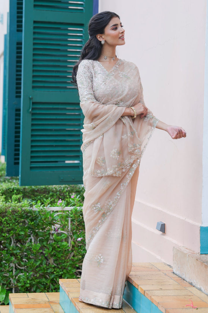 Woman in a beige saree standing outdoors near a building with green shutters.