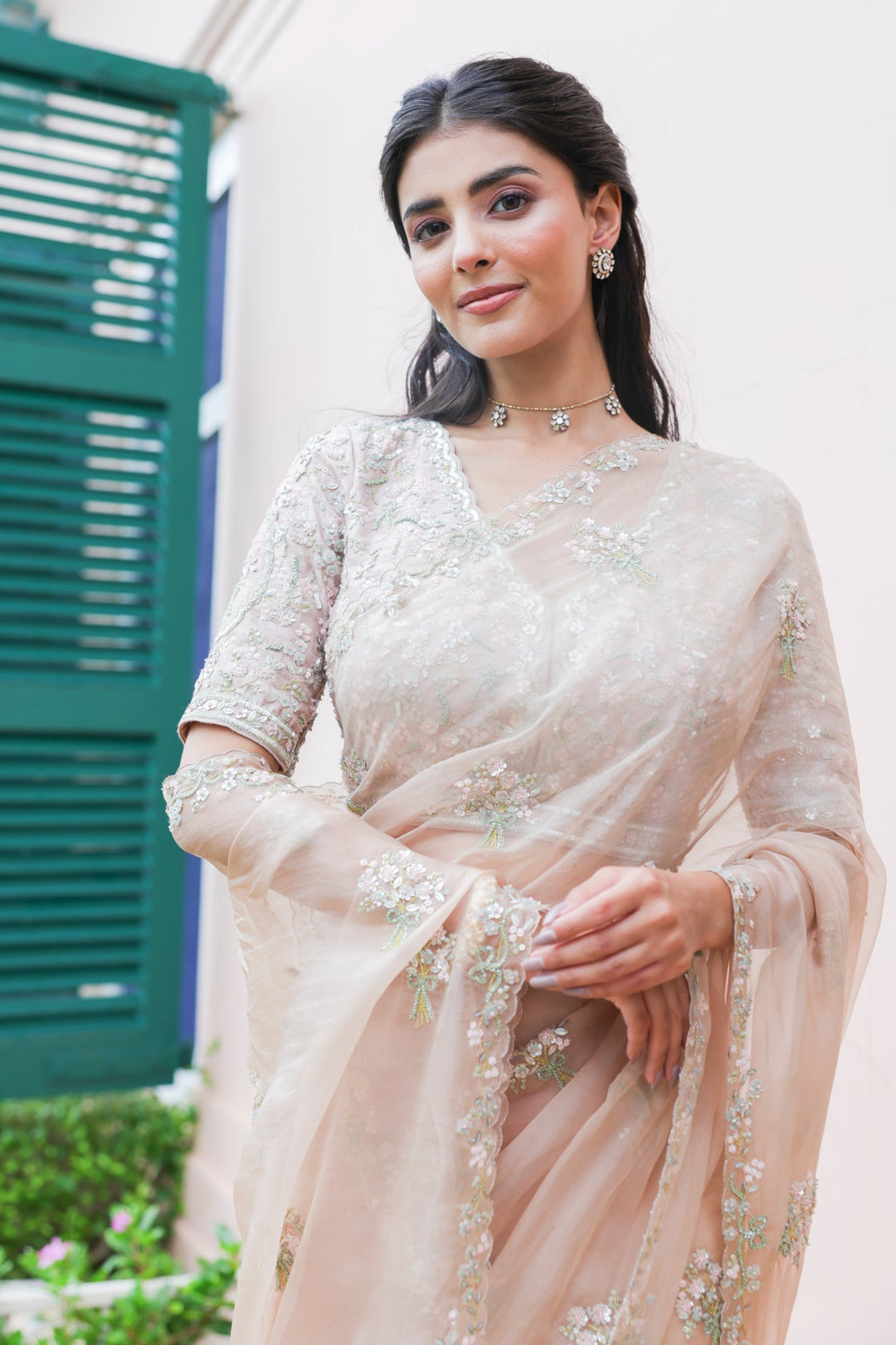 Woman in a light pink saree with floral patterns standing outdoors.