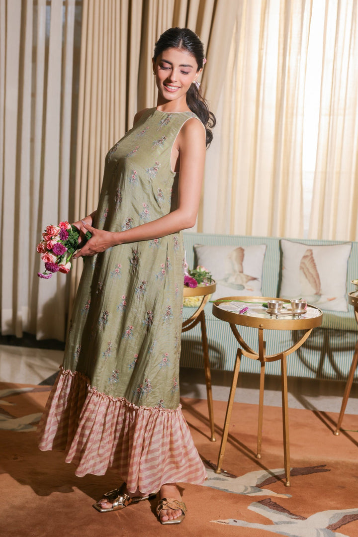 Woman in a green dress holding flowers in a room with curtains and furniture.