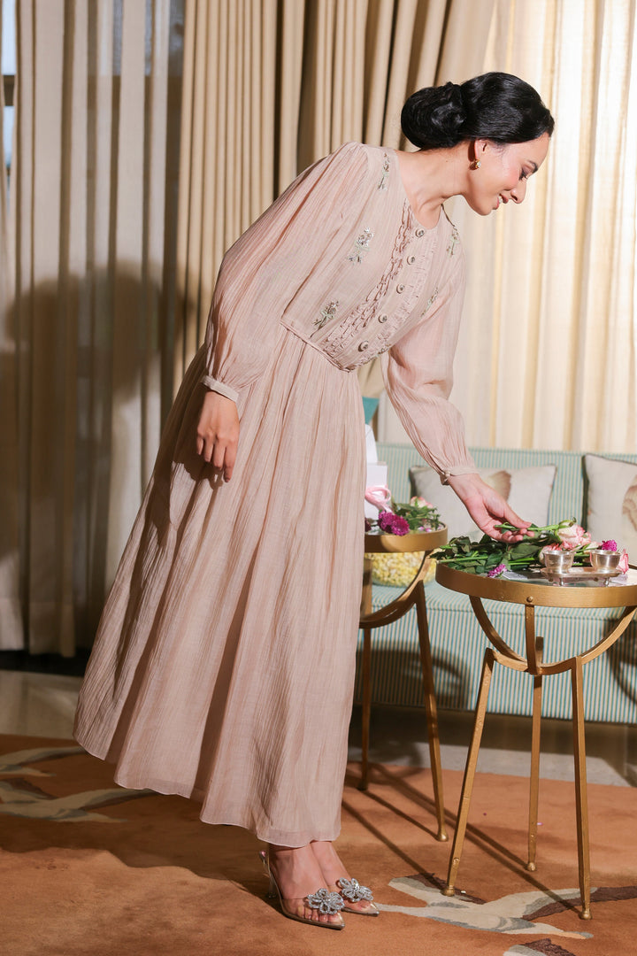 Woman in a long, light-colored dress arranging flowers in a room with curtains and a table.