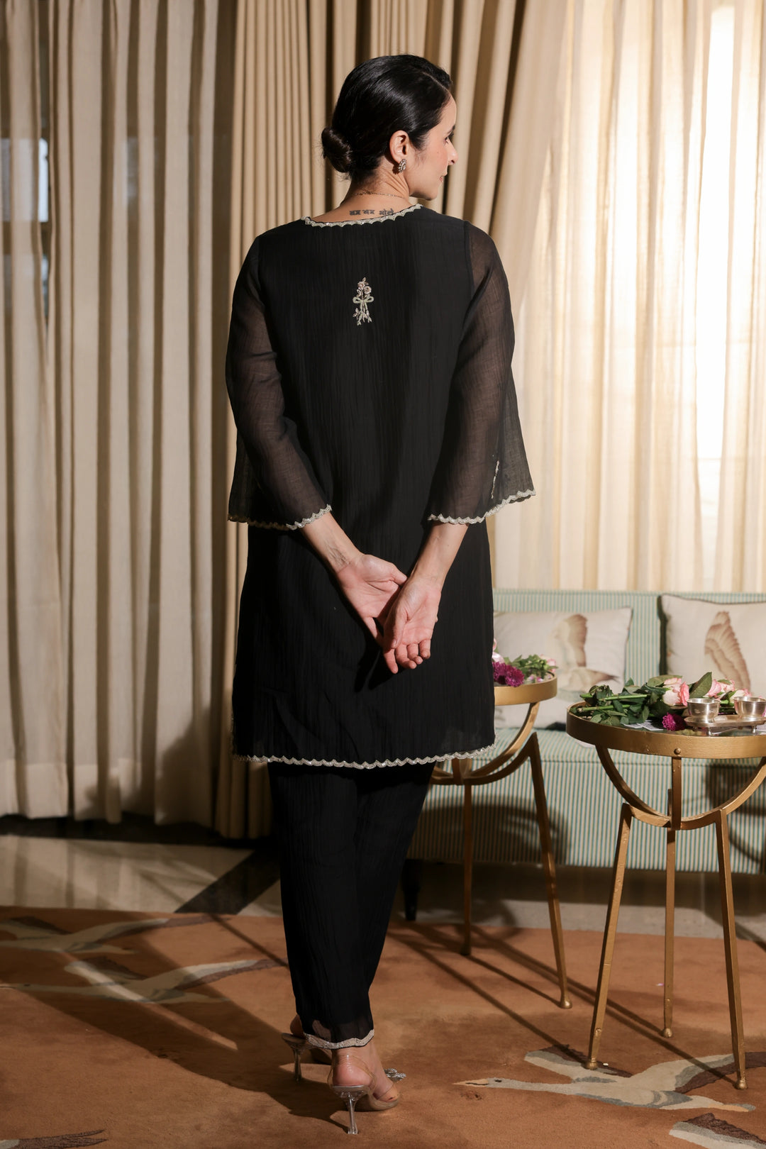 Woman in a black outfit standing in a room with curtains and a table.