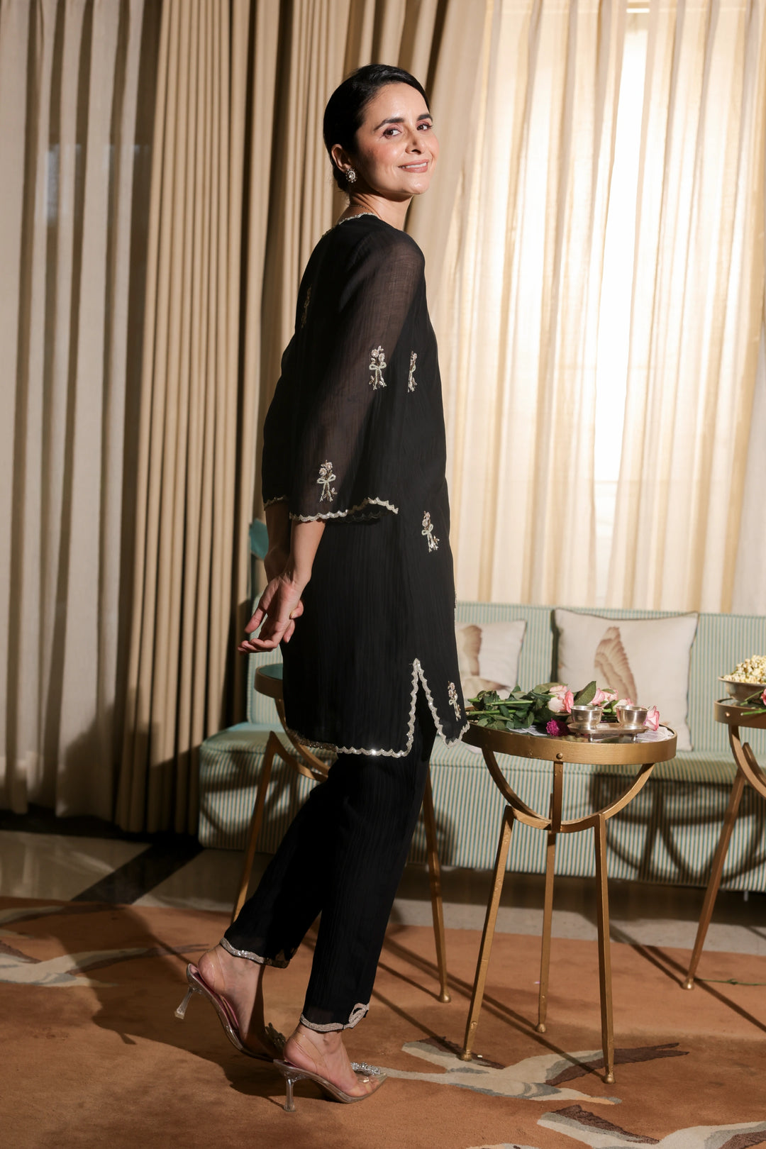 Woman in a black outfit standing in a room with curtains and a table.
