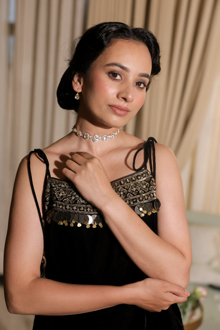 Woman wearing a black dress with gold detailing and a silver necklace against a neutral background