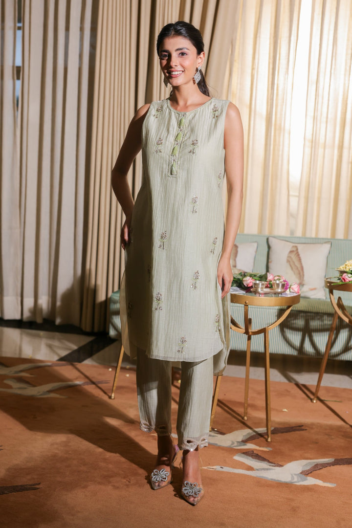 Woman in a light green outfit standing in a room with curtains and a table.
