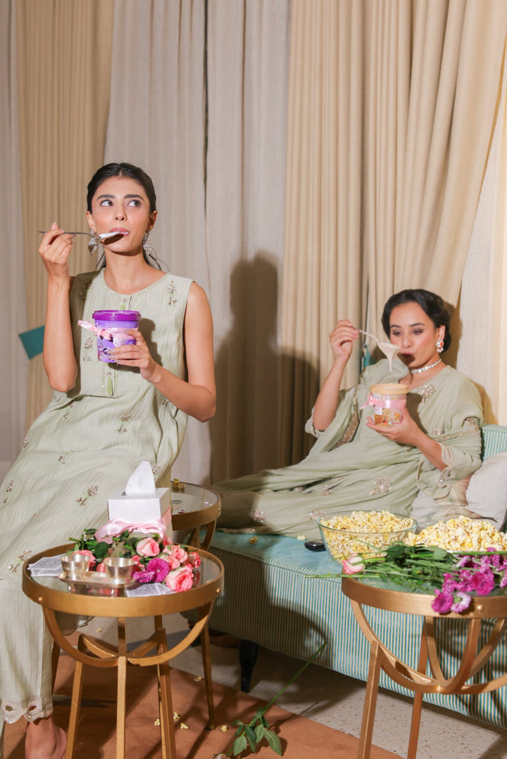 Two women in light green dresses sitting on a couch with snacks and drinks.