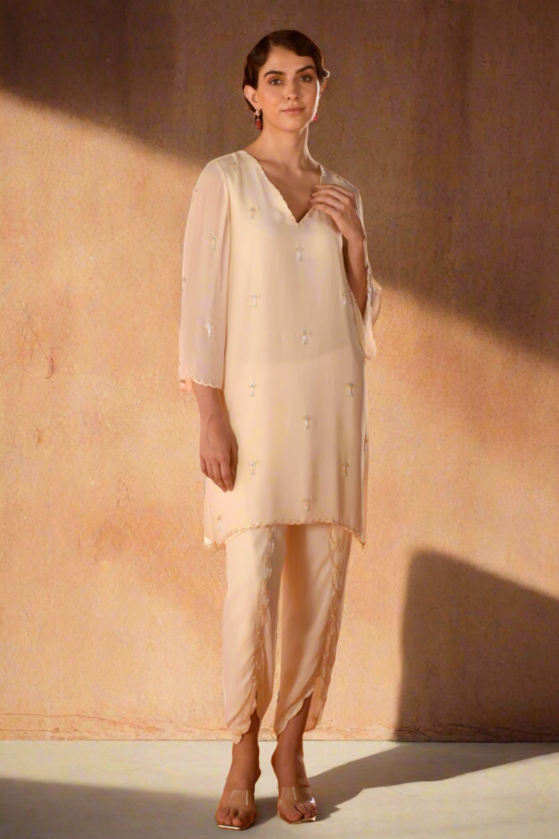 A female model wearing a beige georgette co-ord set with bow embellishments, posing against a brown background.