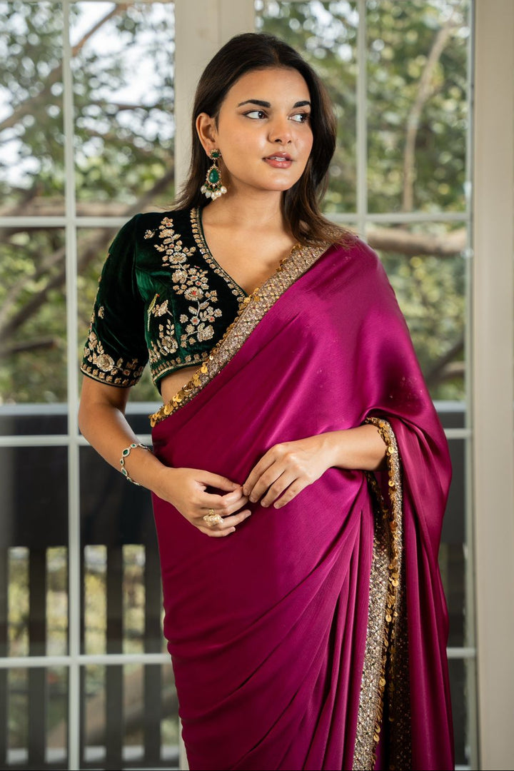 A woman standing in a brightly lit room wearing a traditional purple saree with floral embellishments and a matching velvet blouse.