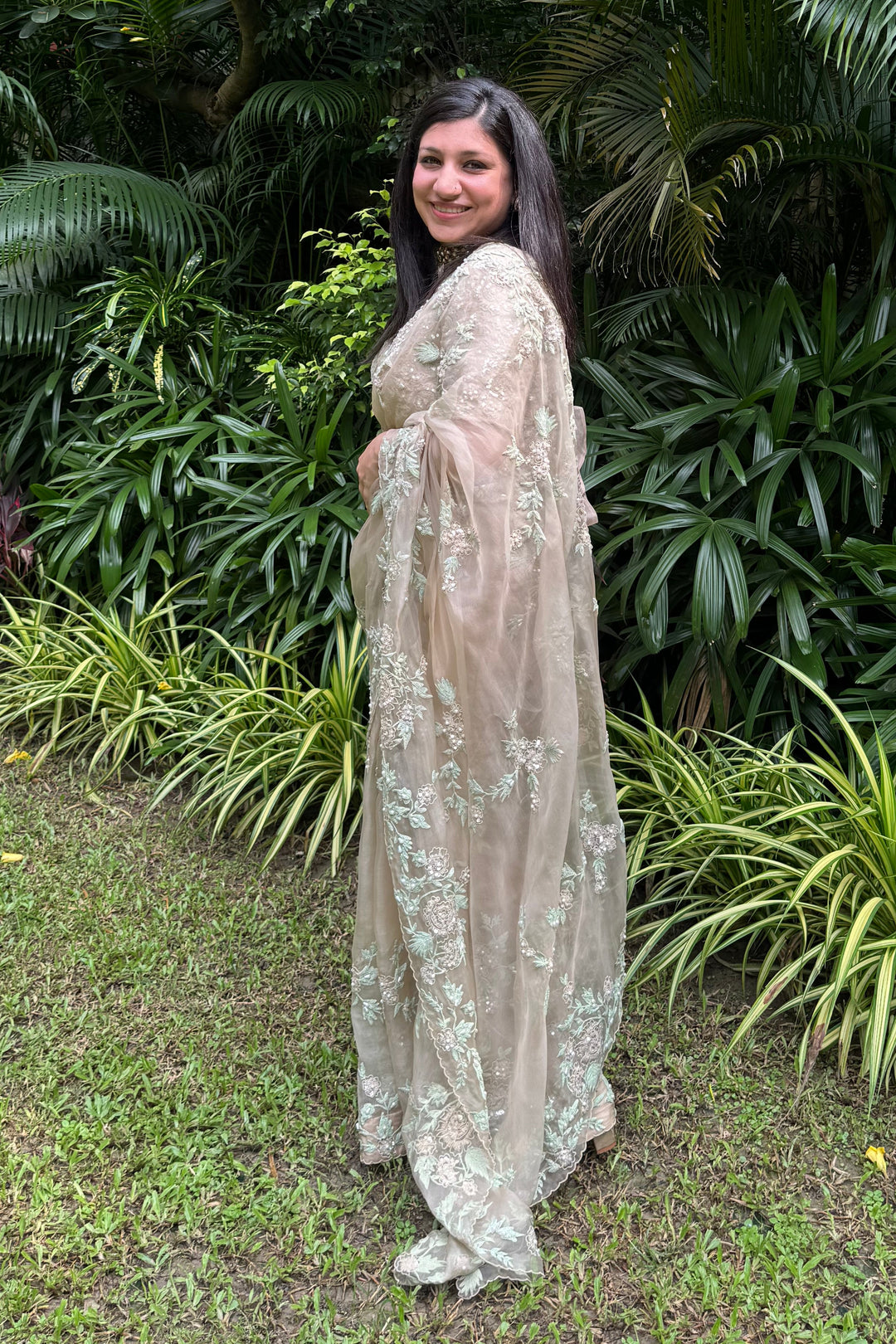 Woman in a light-colored saree standing in a lush green garden
