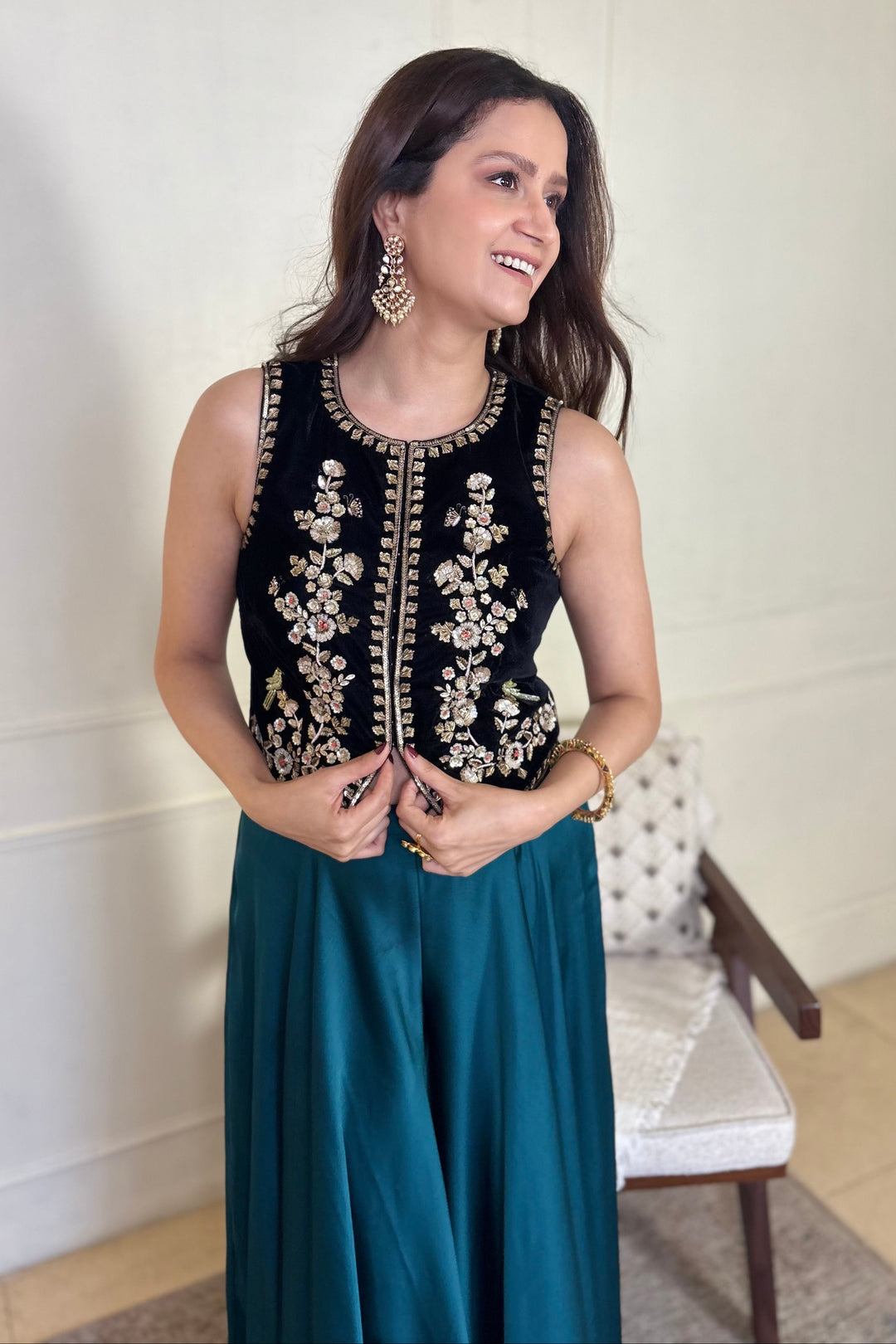 Woman wearing a black embroidered top and teal skirt indoors.