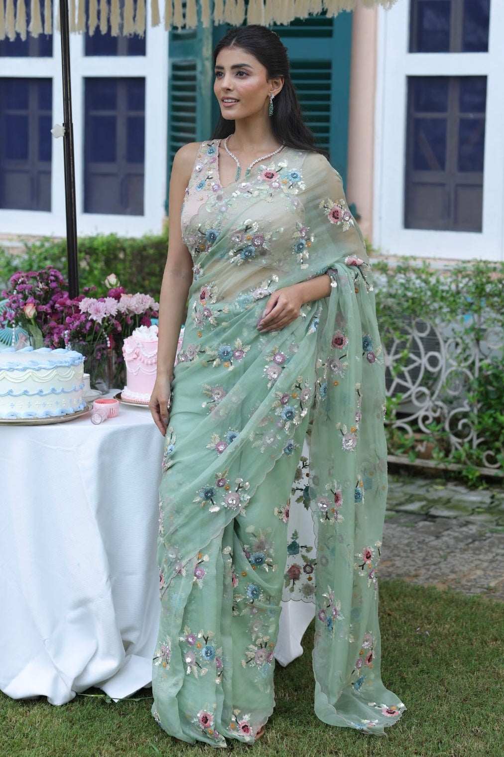 Woman in a light green saree with floral patterns standing outdoors under an umbrella.