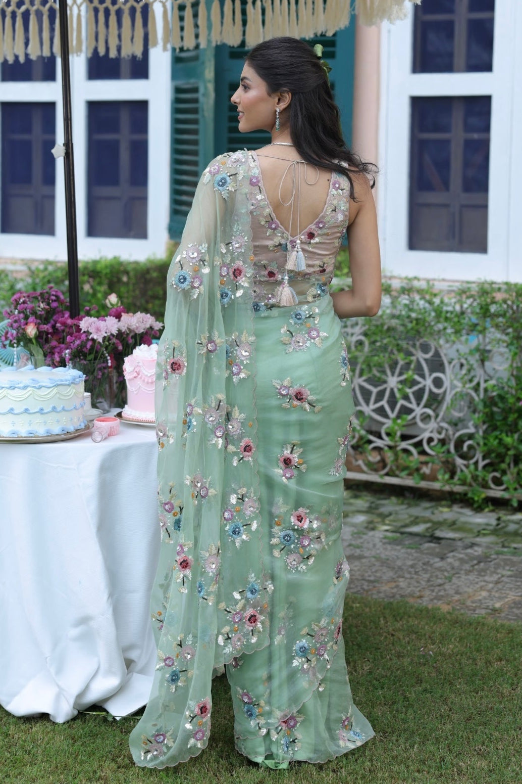 Woman in a light green saree with floral patterns standing outdoors under a decorative umbrella.