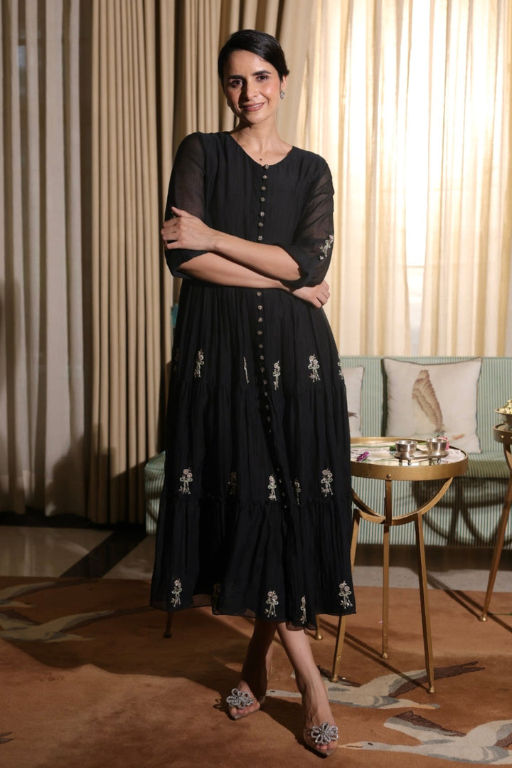 Woman in a black dress standing in a room with curtains and a table.