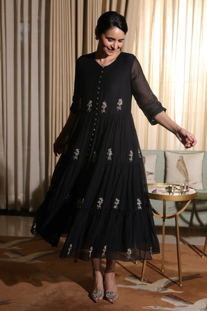 Woman in a black dress with floral embroidery standing in a room with curtains and a table.