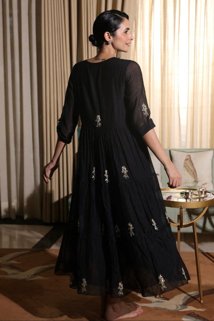 Woman in a black dress with floral embroidery standing in a room with beige curtains.