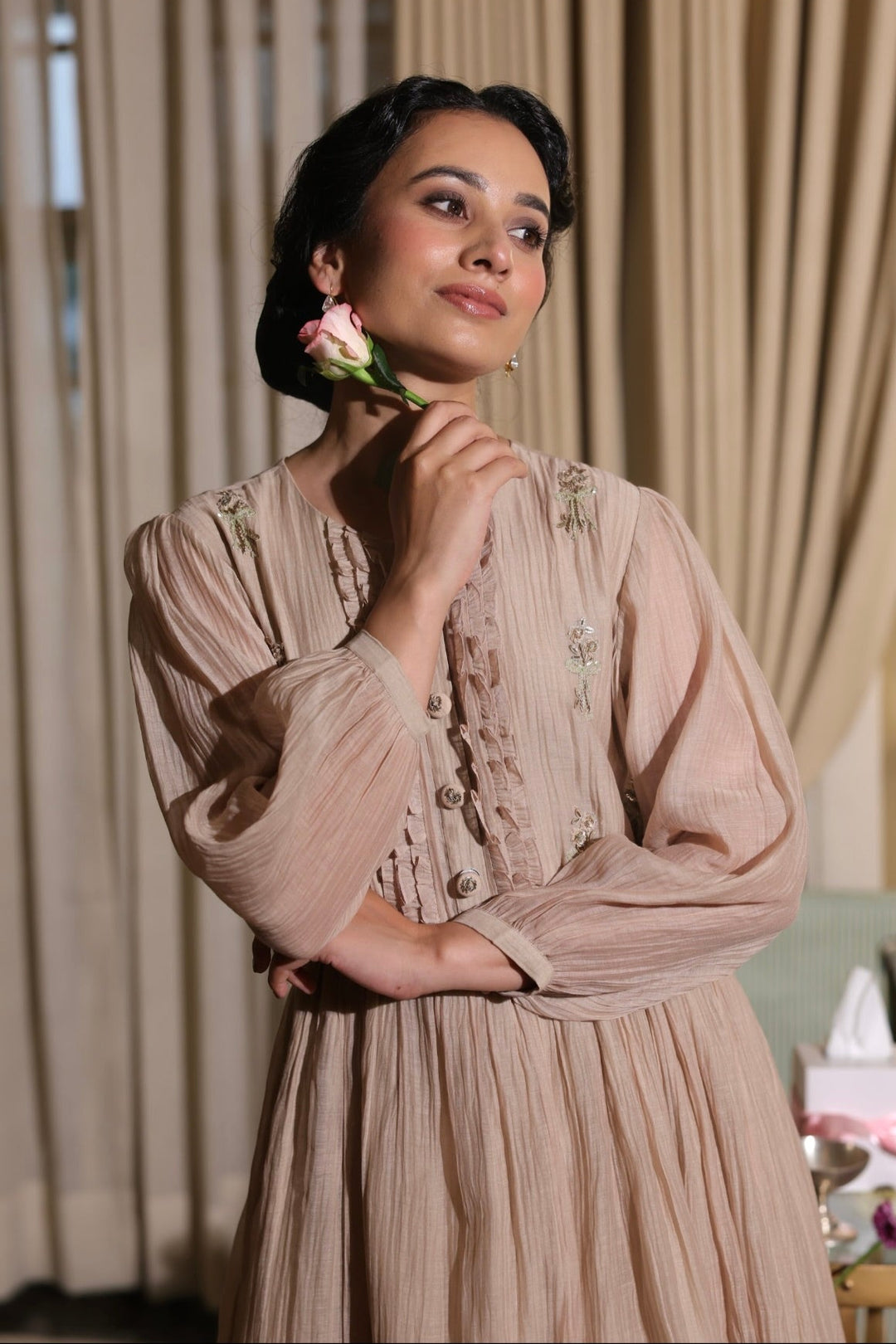 Woman in a vintage-style dress holding a flower in front of beige curtains.