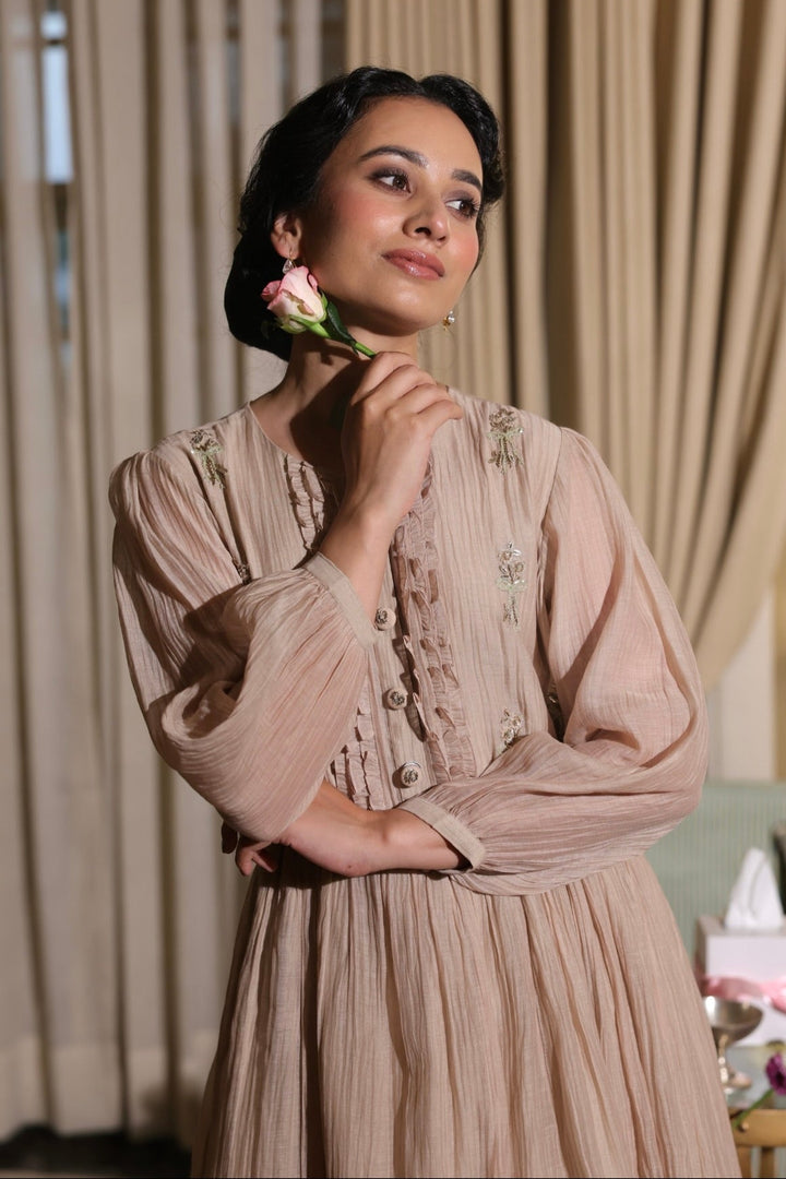 Woman in a vintage-style dress holding a flower in front of beige curtains.