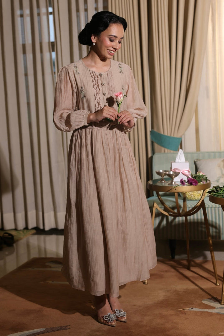 Woman in a long beige dress standing indoors with curtains and furniture in the background