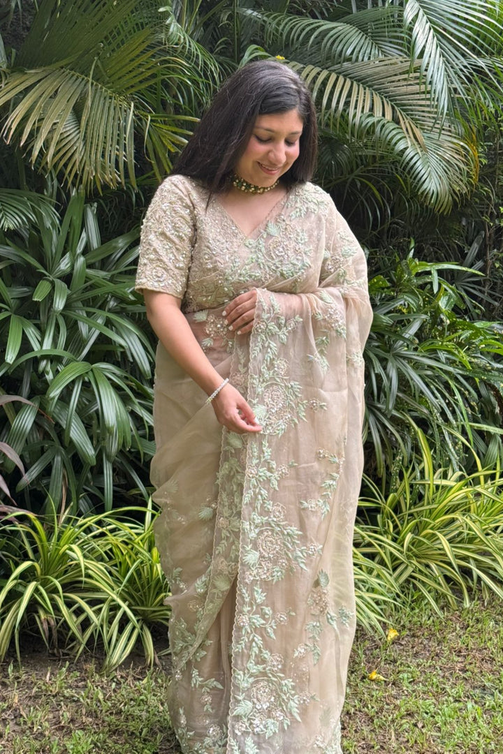 Woman in a beige saree with floral patterns standing in front of green foliage