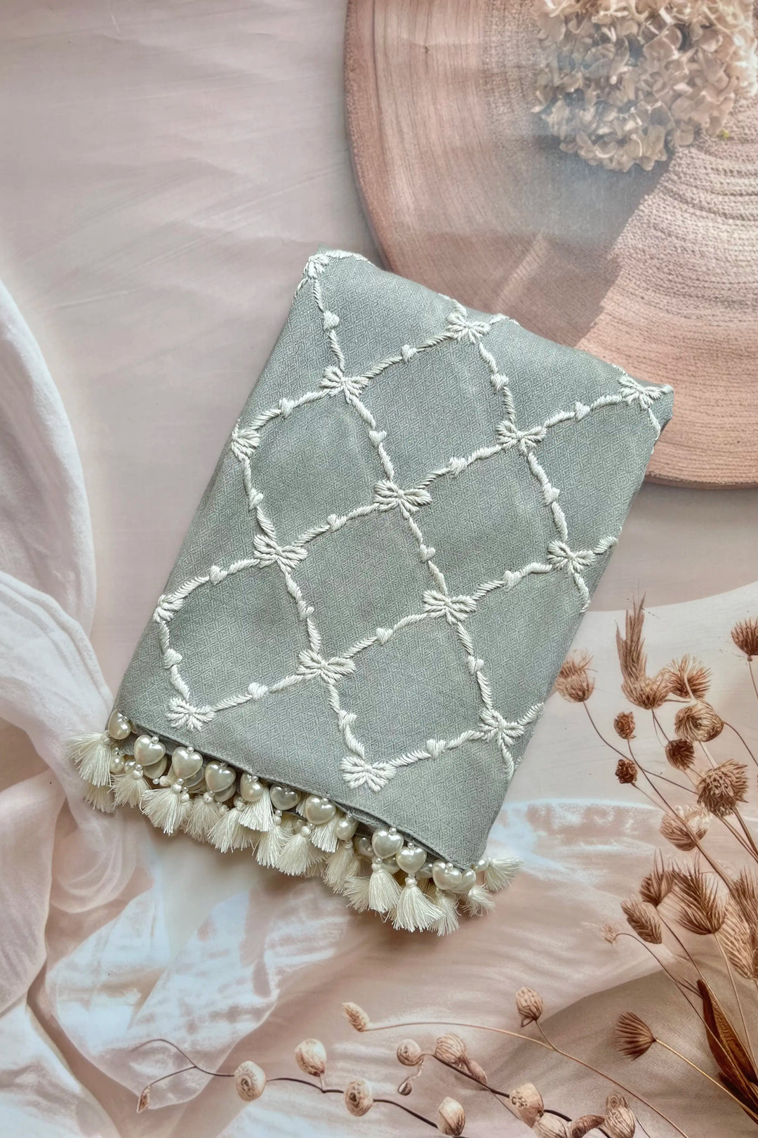 Decorative pillow with white embroidery and tassels on a soft surface with dried flowers.