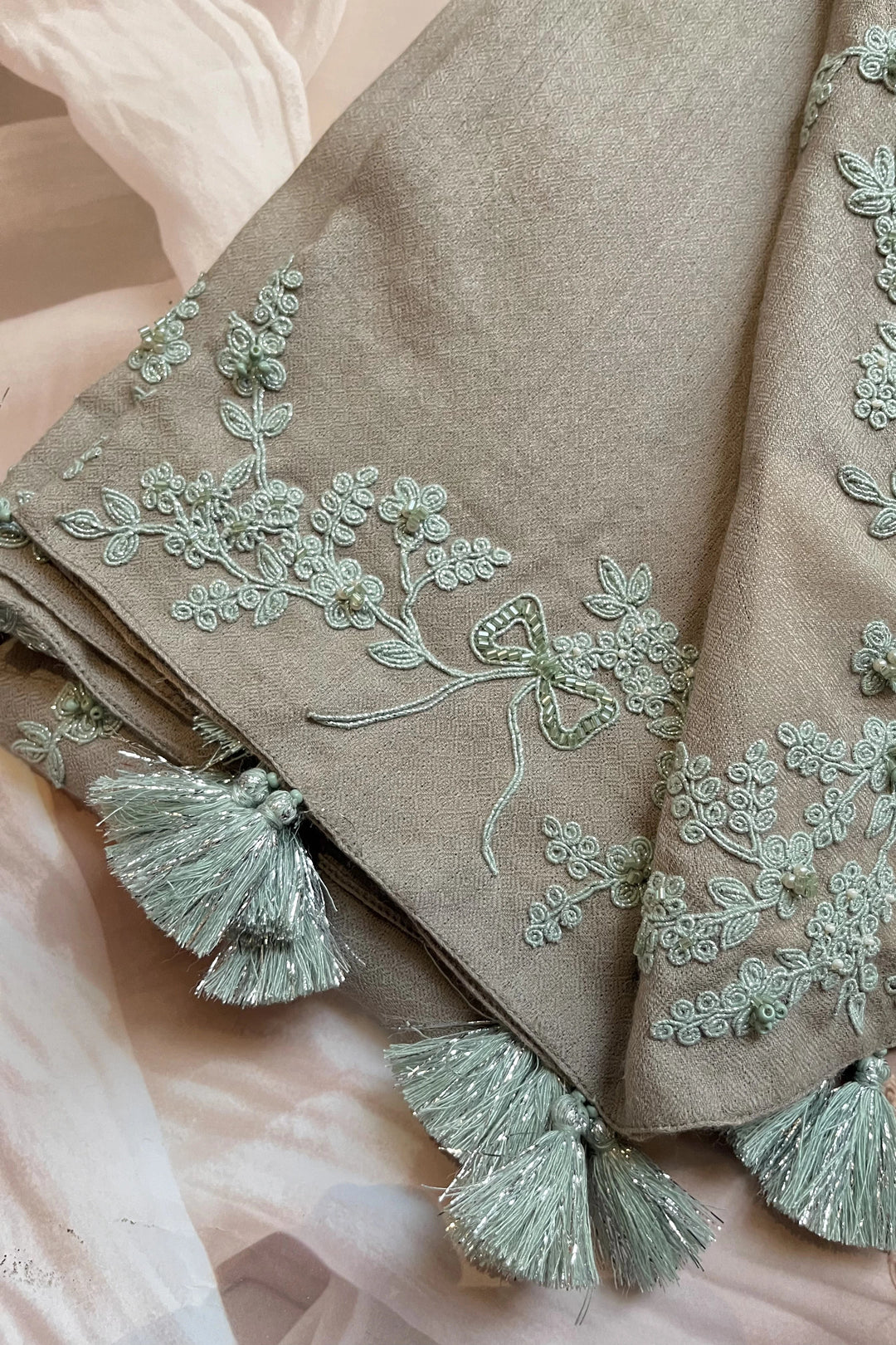 Brown fabric with intricate green embroidery and tassels on a soft pink background
