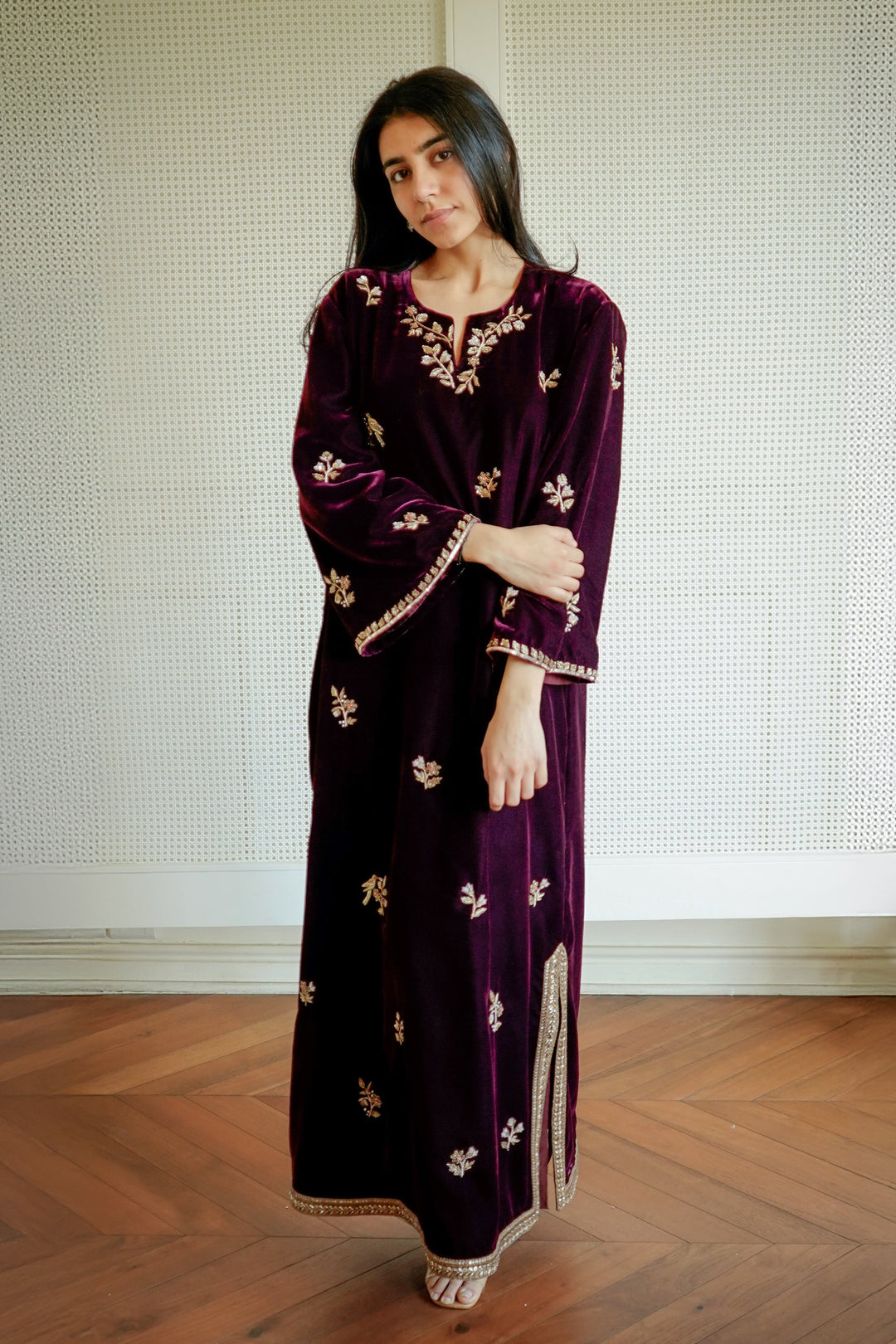 Woman wearing a dark purple dress with gold patterns standing in a room with wooden flooring and a white wall.