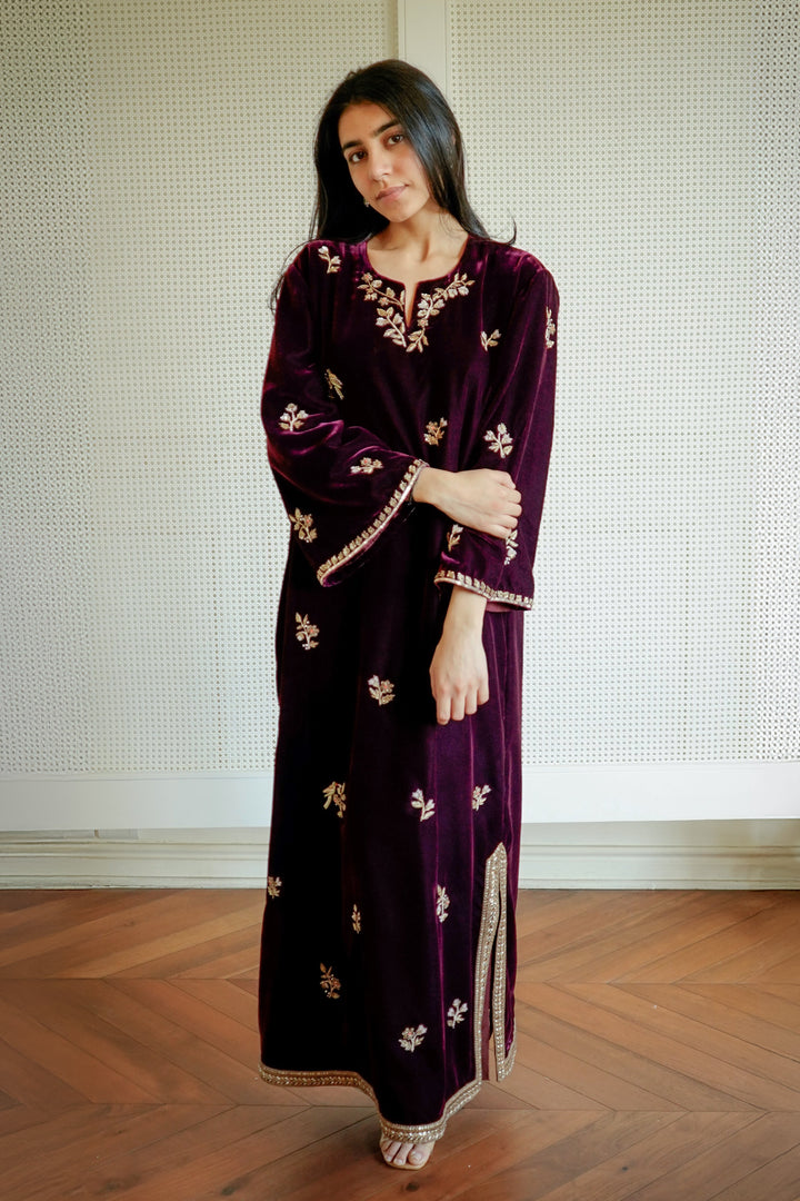 Woman wearing a dark purple dress with gold patterns standing in a room with wooden flooring and a white wall.