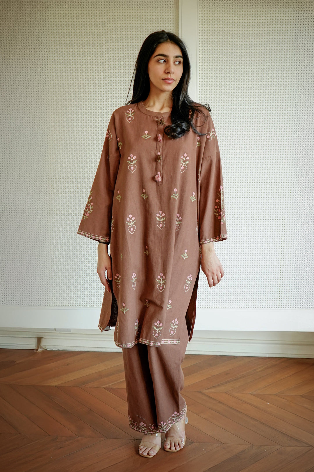Woman wearing a brown embroidered outfit standing on a wooden floor with a white wall background