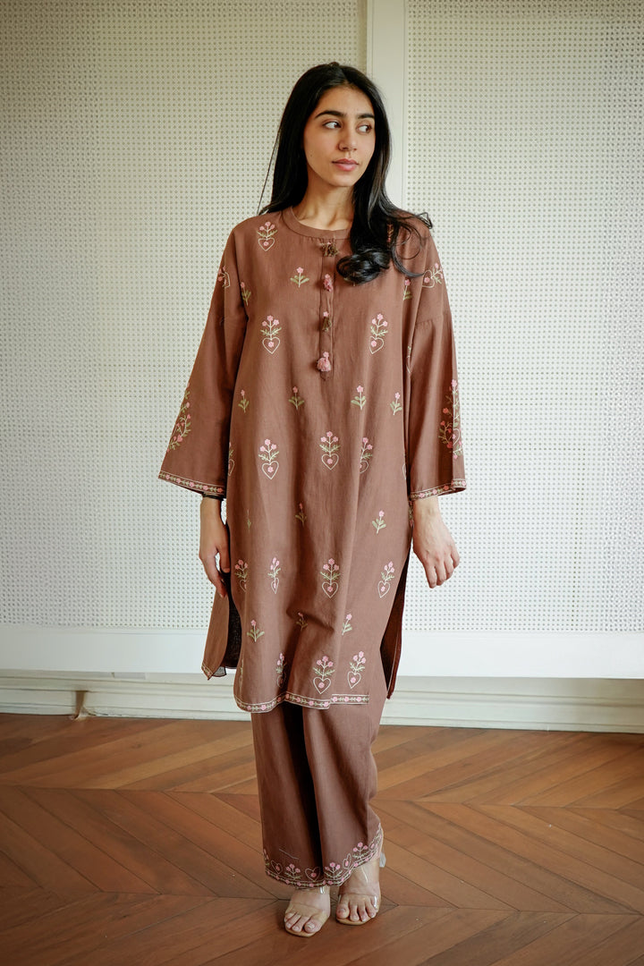 Woman wearing a brown embroidered outfit standing on a wooden floor with a white wall background