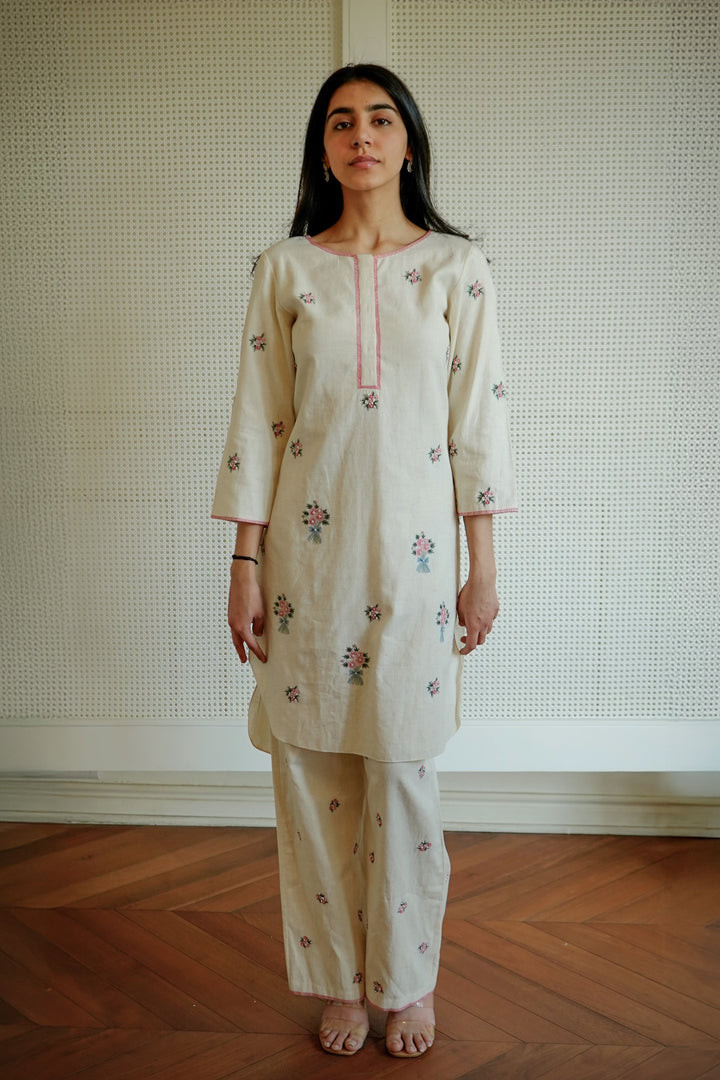 Woman wearing a cream-colored embroidered outfit standing against a textured wall.