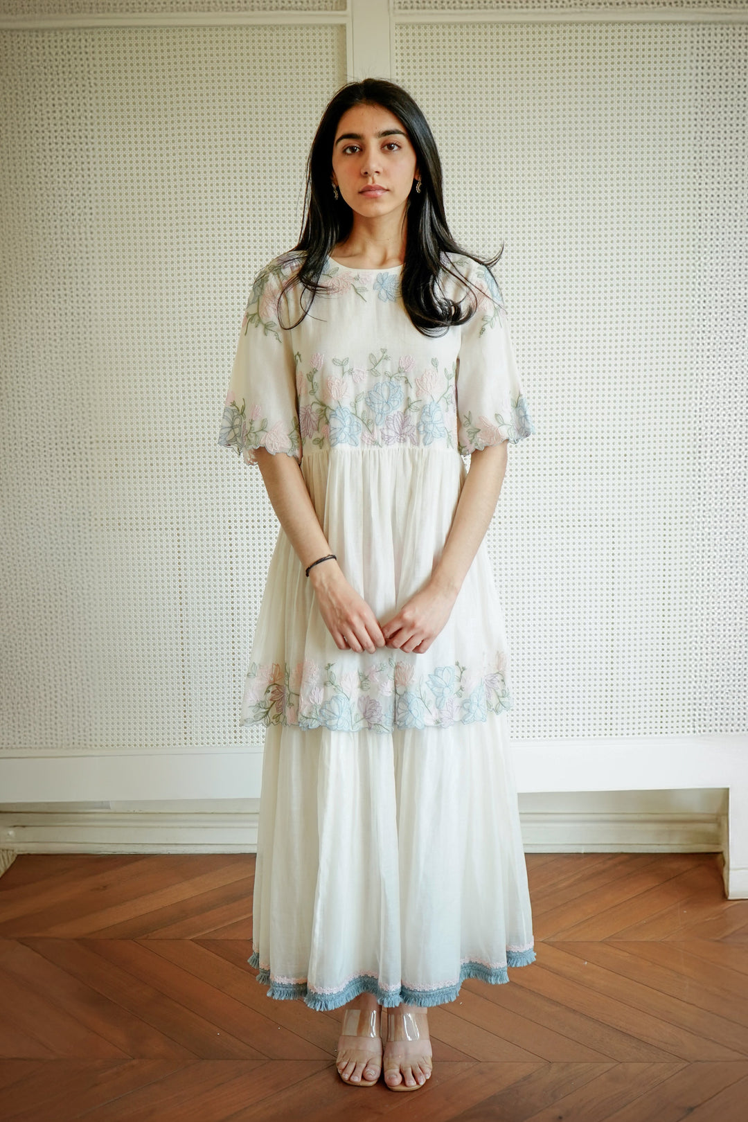 Woman wearing a floral dress standing in a room with wooden flooring and a textured wall.