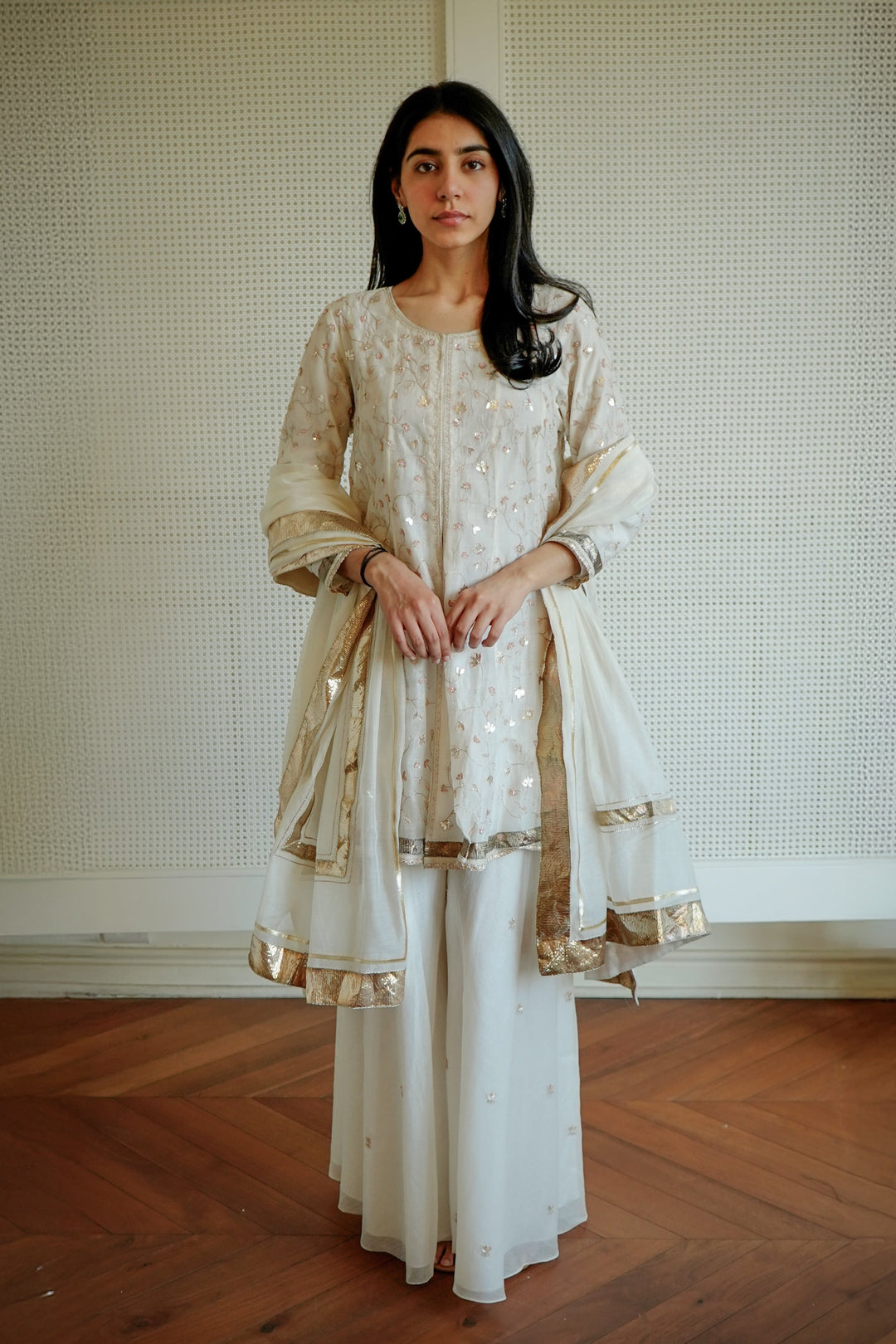 Woman wearing a traditional outfit with gold and white patterns against a neutral background.