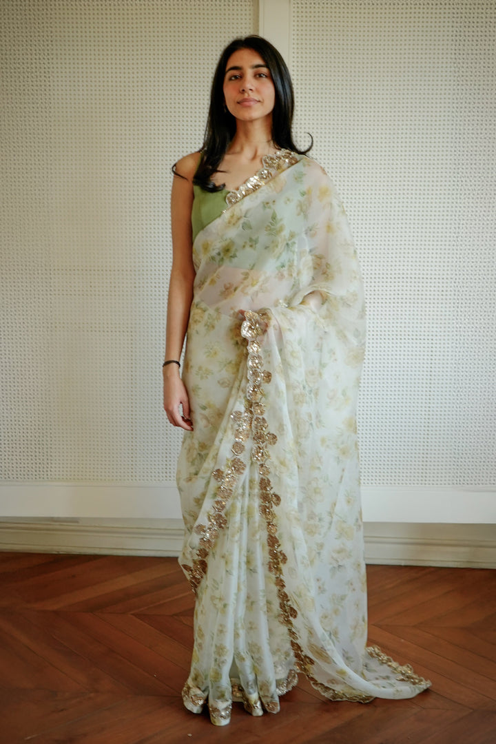 Woman wearing a white saree with gold patterns in a room with wooden flooring and a white wall.