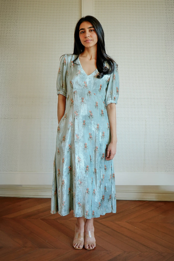 Woman wearing a light blue floral dress standing against a neutral wall.