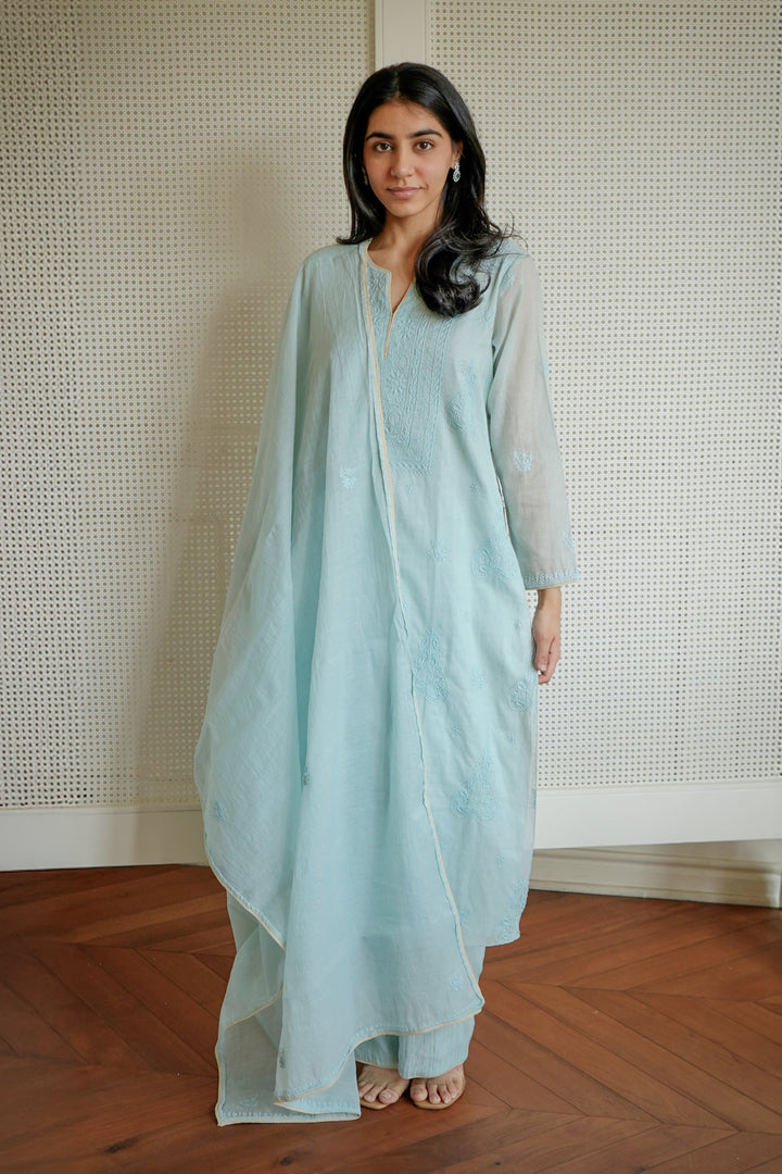 Woman wearing a light blue dress standing in a room with wooden flooring and a textured wall.