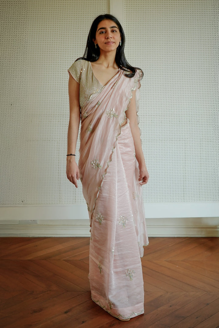 Woman in a light pink saree standing in a room with wooden flooring and a textured wall.