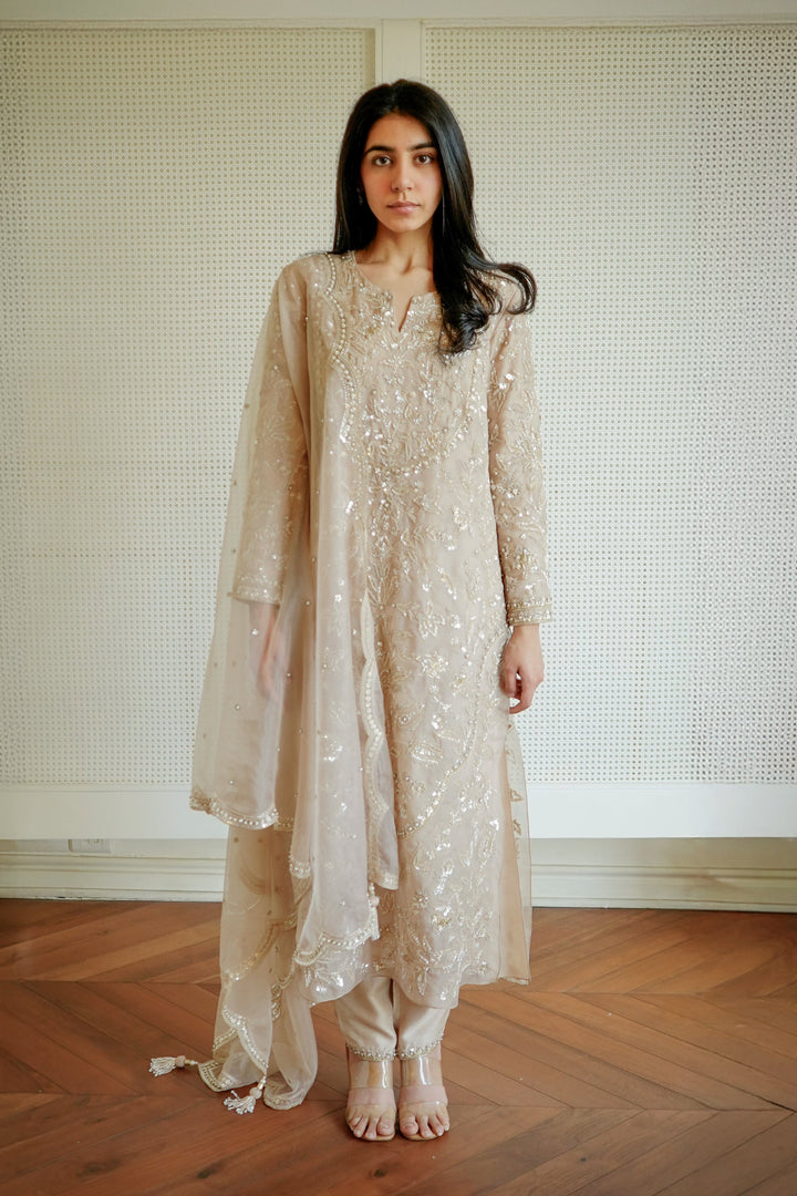 Woman wearing a beige embroidered dress standing in a room with wooden flooring and a textured wall.