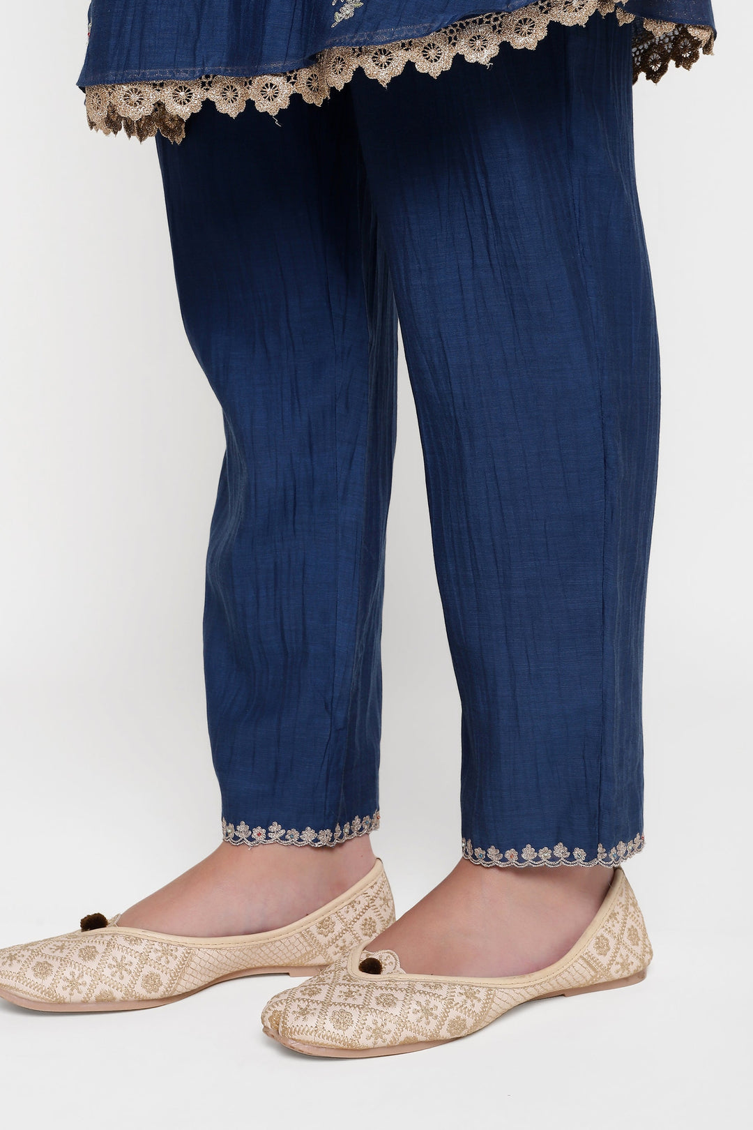 Blue pants with white lace trim worn by a person, paired with gold embroidered flats on a white background.