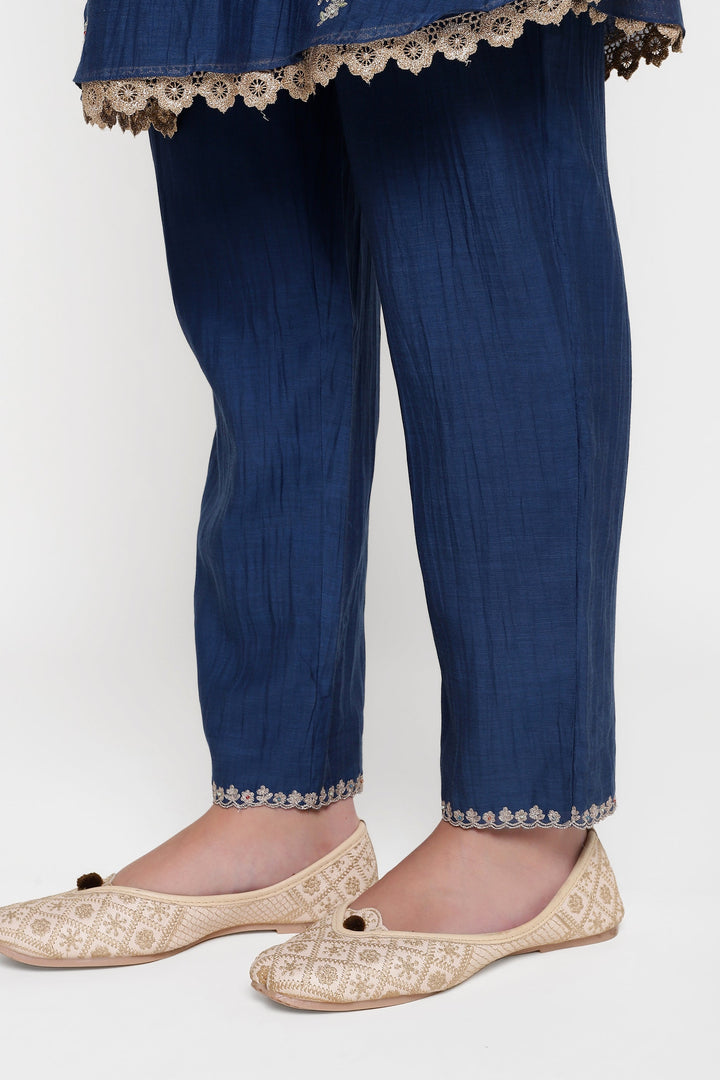 Blue pants with white lace trim worn by a person, paired with gold embroidered flats on a white background.