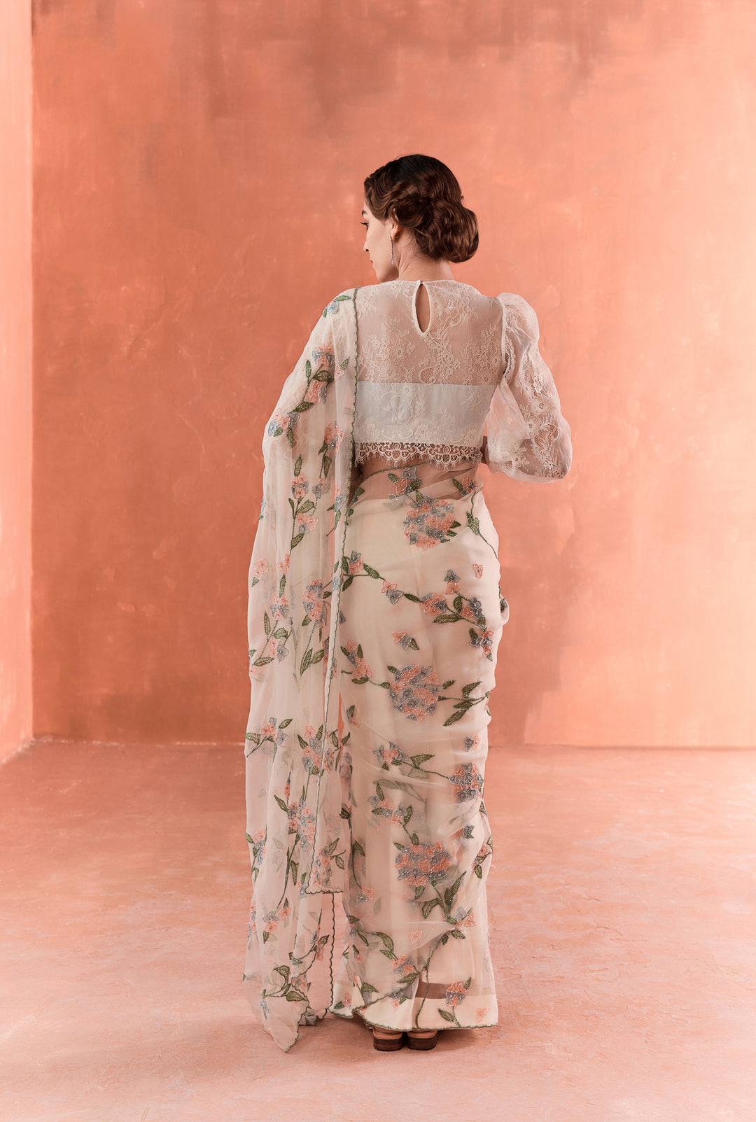 Woman wearing a floral dress with a sheer dupatta against a pink wall.