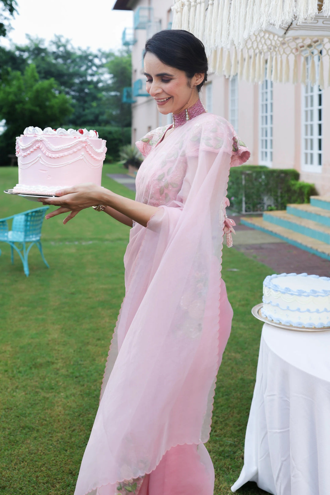 Woman in a pink dress holding a pink cake outdoors with a house in the background