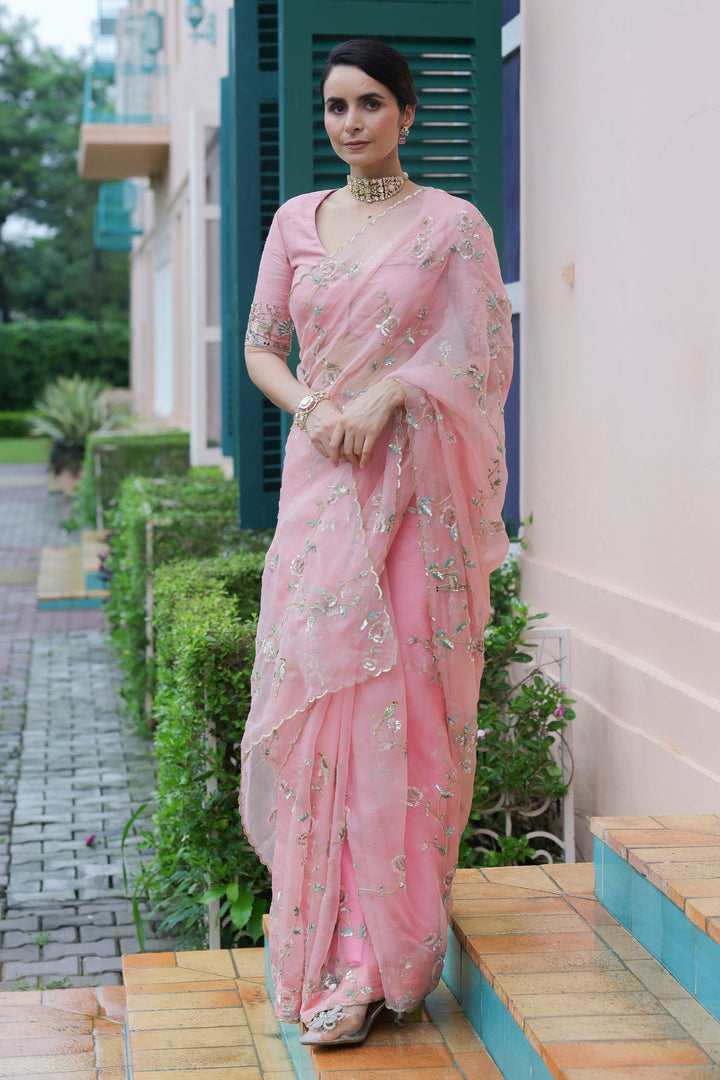Woman in a pink saree standing on a wooden platform with a building and greenery in the background